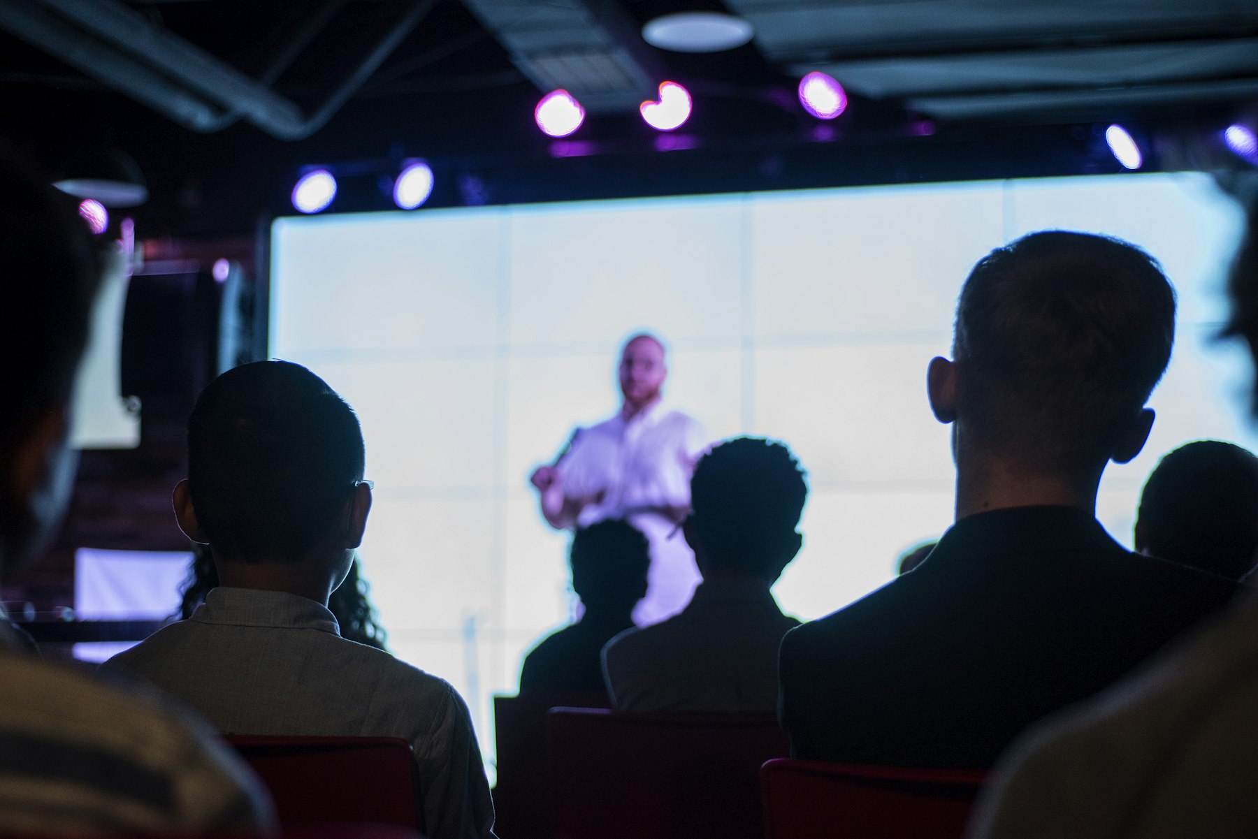 Audience seen from behind watching a speaker on a very large bright screen in a dark hall with stage lights.