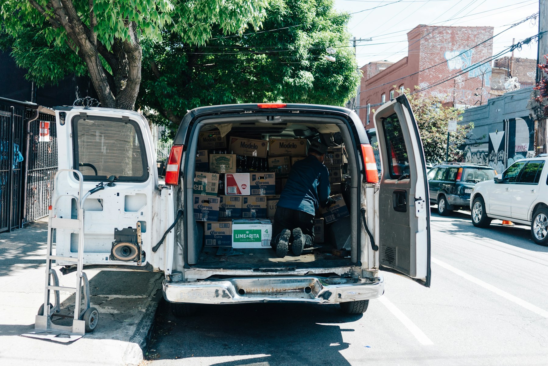 A man kneels in the open back of a white cargo van stacking boxes on a sunny street, hand truck beside it.
