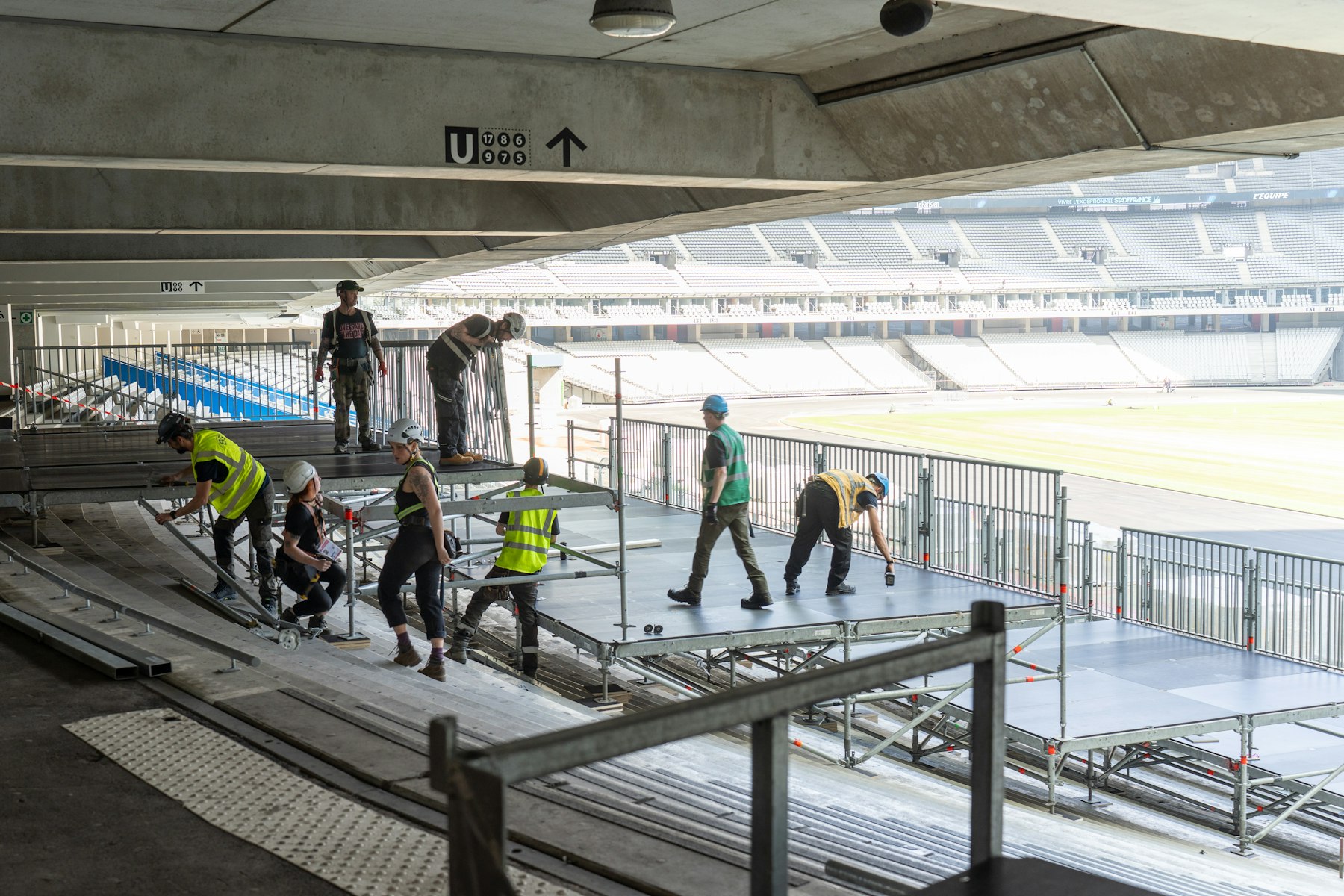 Workers in hi-vis vests and hard hats assemble a modular platform stage on stadium stands.