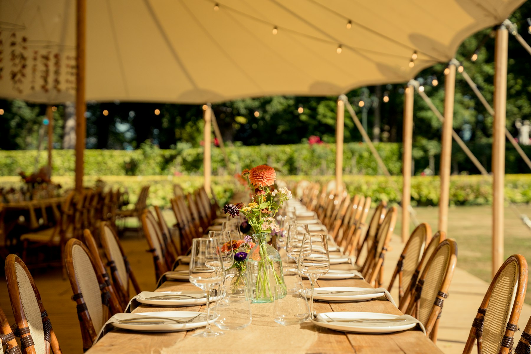 Long dining table with plates and glasses under a cream tent canopy hung with string lights in a garden.